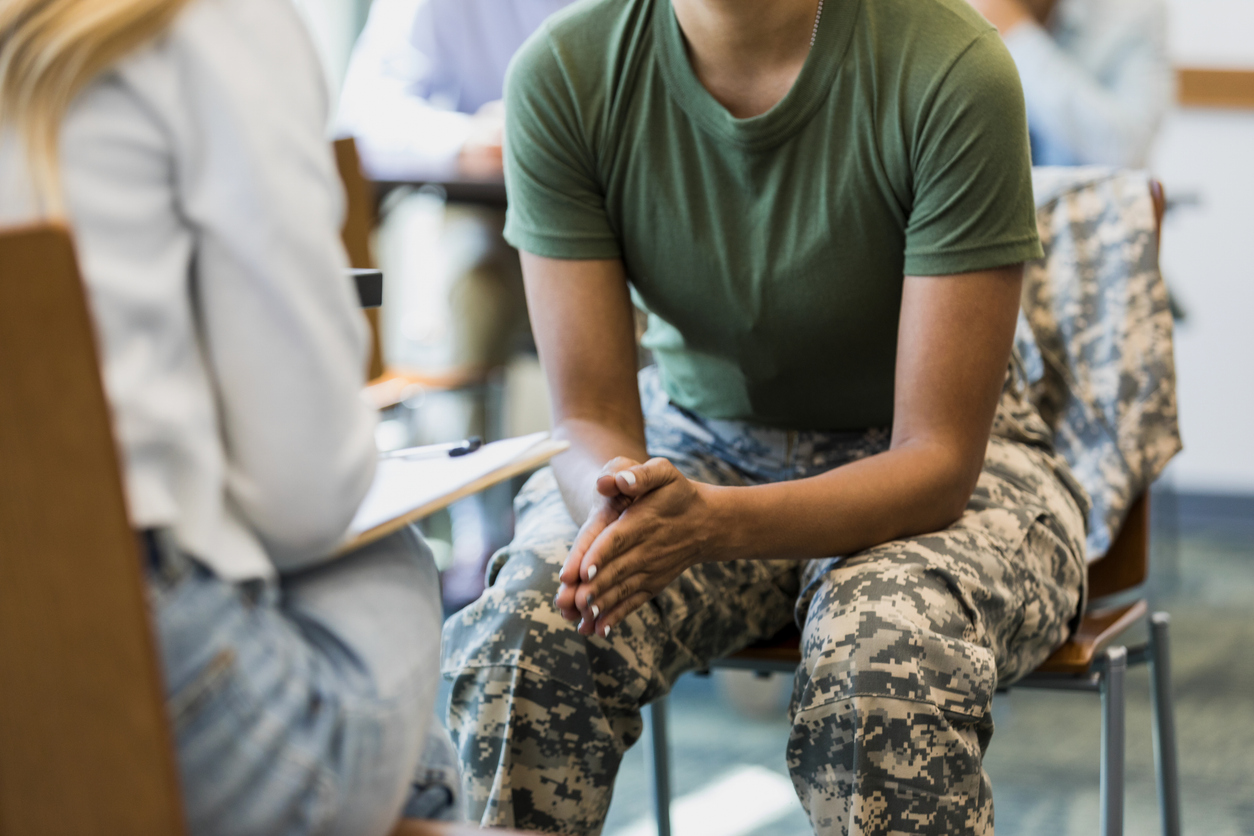 A close up photo of an unrecognizable mid adult female soldier as she puts her hands together and leans forward in her seat. She is talking with an unrecognizable female counselor.