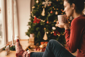 Young woman drinking tea by the Christmas tree, looking through window.