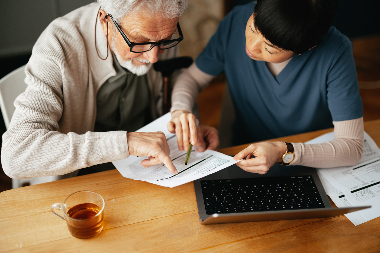 High angle view of a Japanese female caregiver doing home finance online on a computer together with her worried elderly patient at his home.