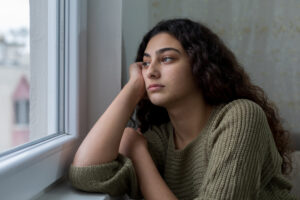 A sad teenage girl looking out a window from a darker room